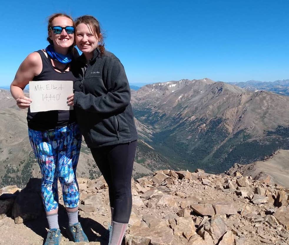 Two women standing atop a mountain holding a sign that reads Mt. Elbert 14,440'