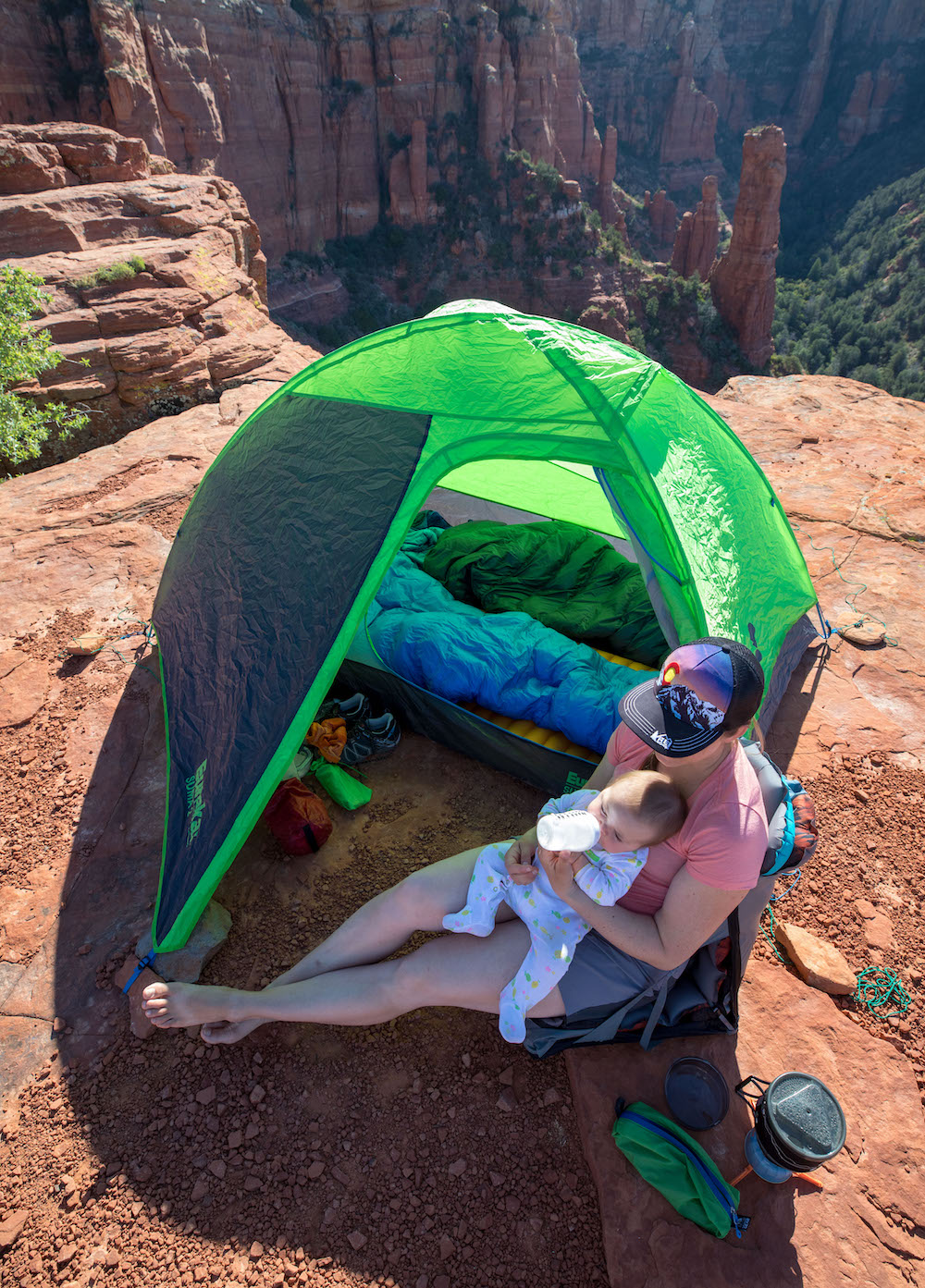 Woman feeding baby with a bottle in front of a tent with red rock pinnacle formations behind her. 