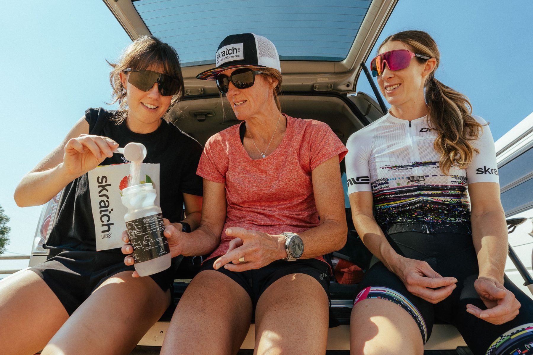 Three women in athletic gear sit on the open tailgate of a car. One woman scoops powder from a Skratch Labs bag into a sports bottle while the others watch and smile. They all wear sunglasses and cycling clothes. Darby Communications PR + Digital Marketing