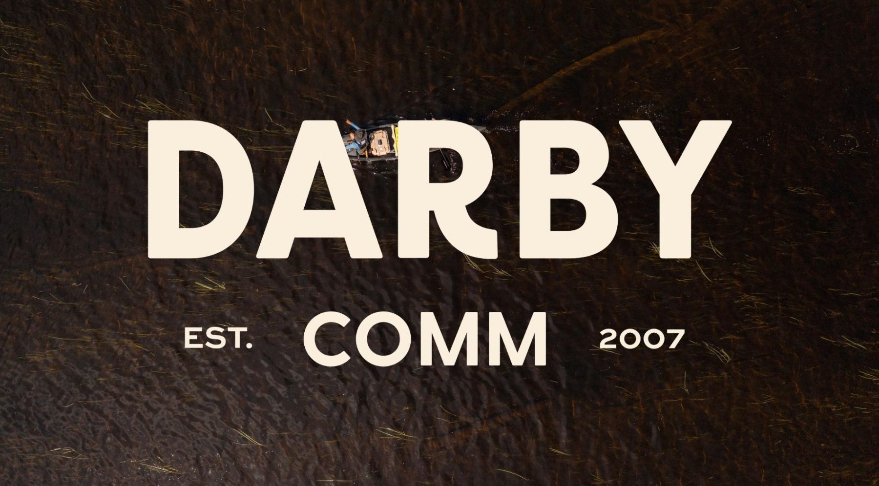 Aerial view of a person in a canoe on dark water, overlaid with large beige text reading DARBY and smaller text below reading EST. COMM 2007. Darby Communications PR + Digital Marketing