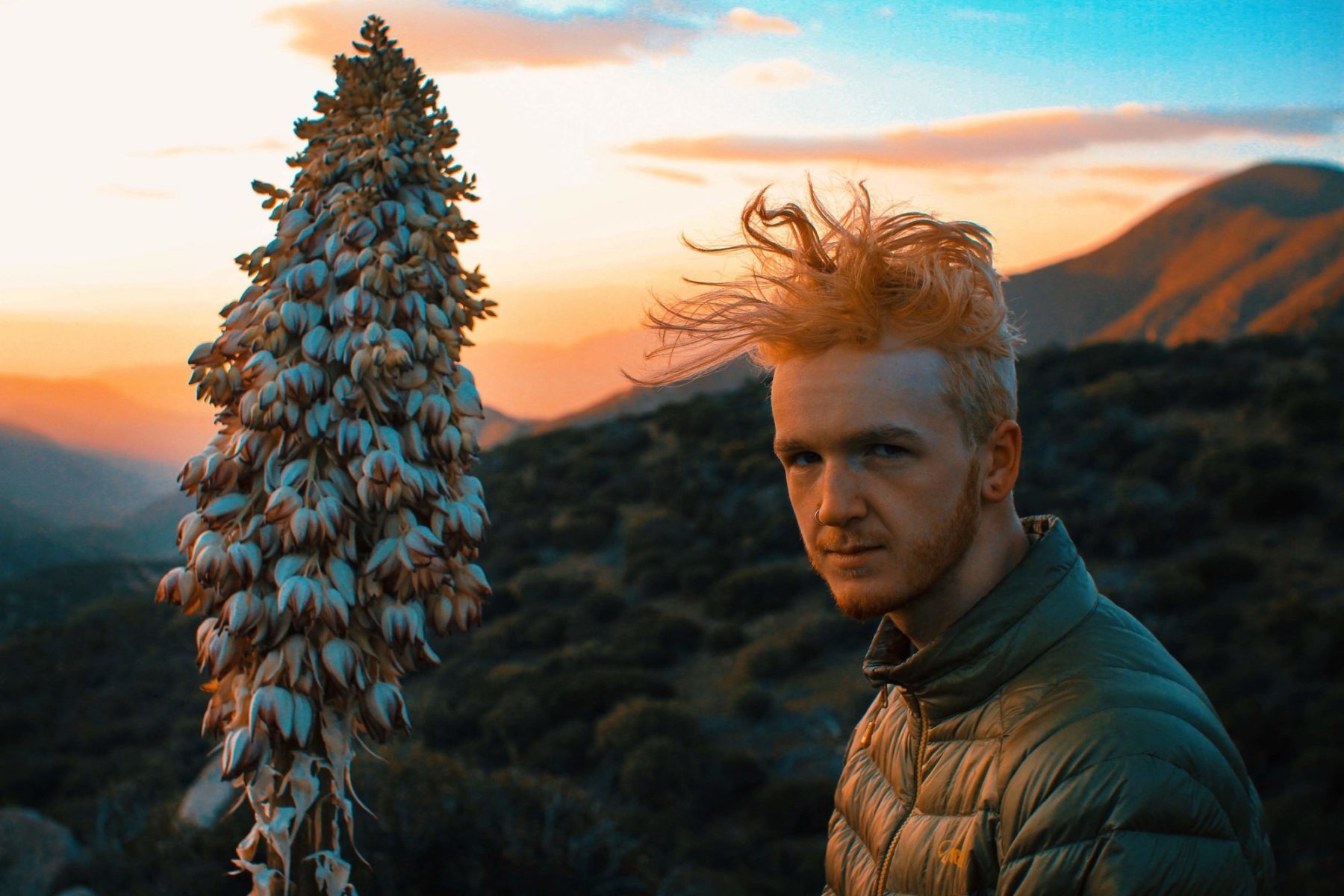 A man with pinkish colored hair stands in front of mountains at sunrise or sunset