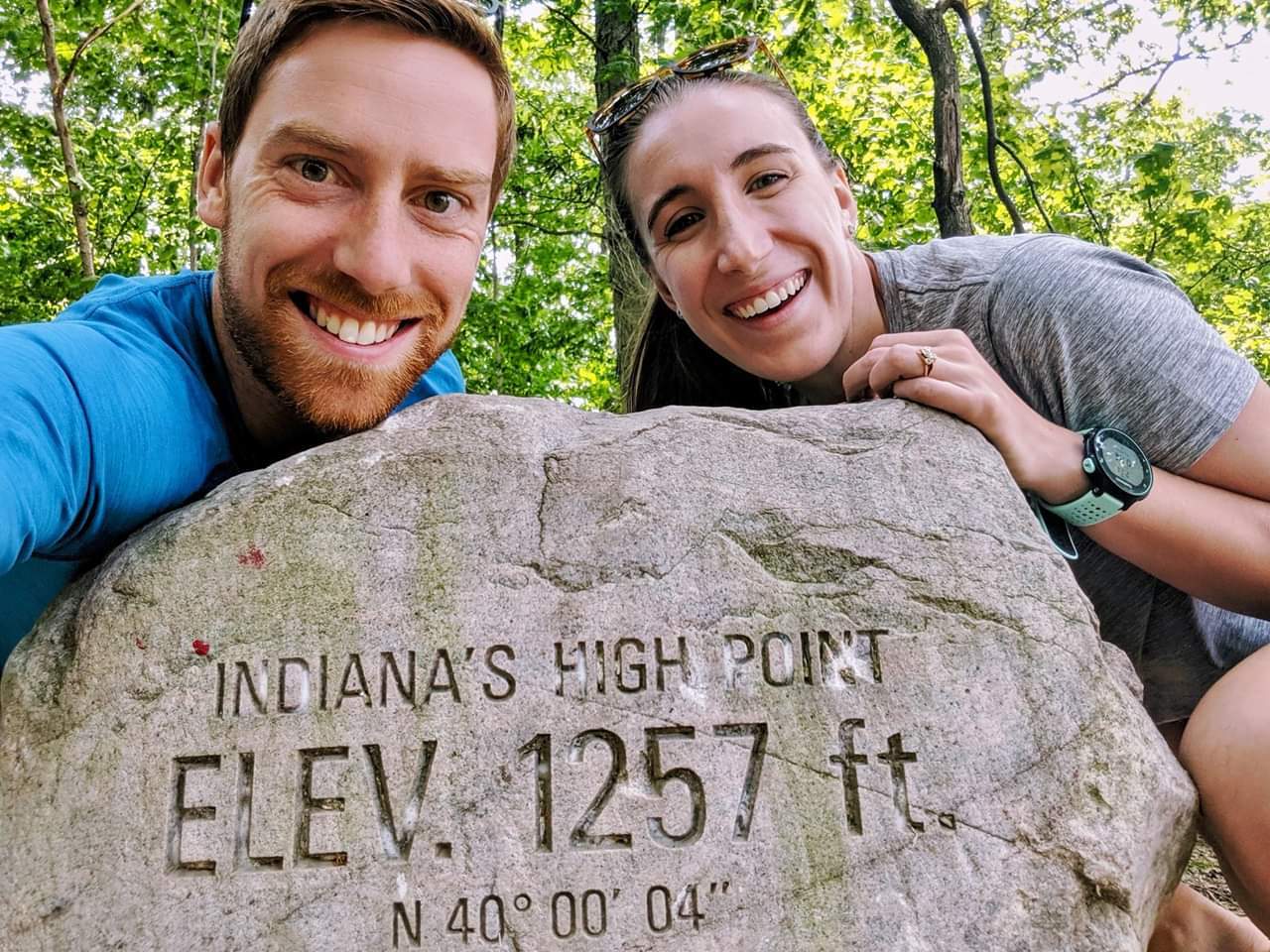A man and woman pose for a picture with a rock that says Indiana's High Point Elev. 1257 ft.
