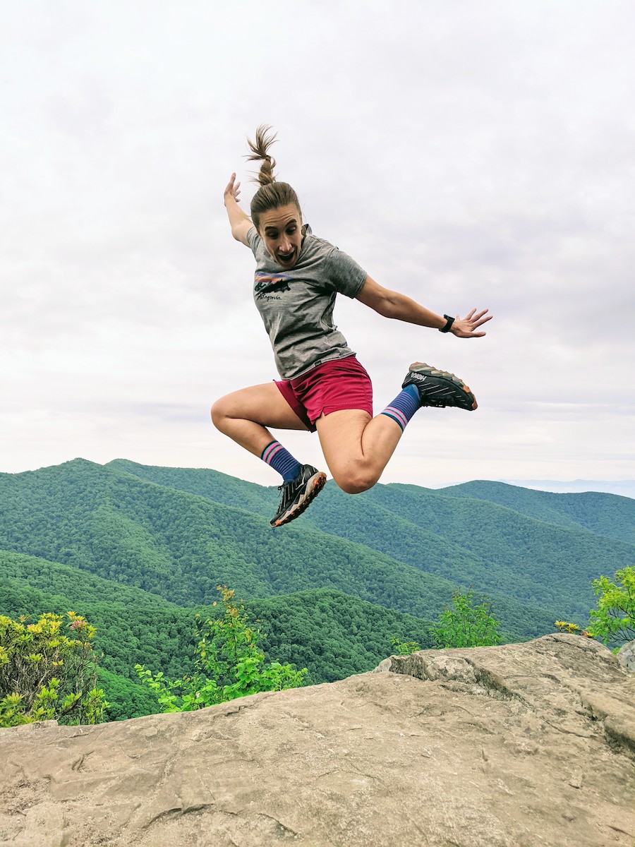 Abby Harris jumping in air in mountains with rock beneath her and green mountains behind her. 
