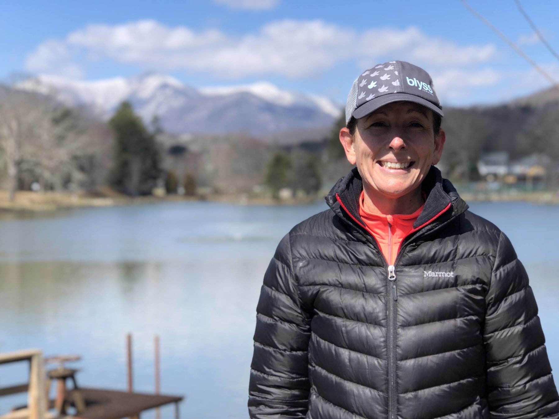 A woman in a trucker hat and puffy jacket in front of a pond with snowcapped mountains in the background. 
