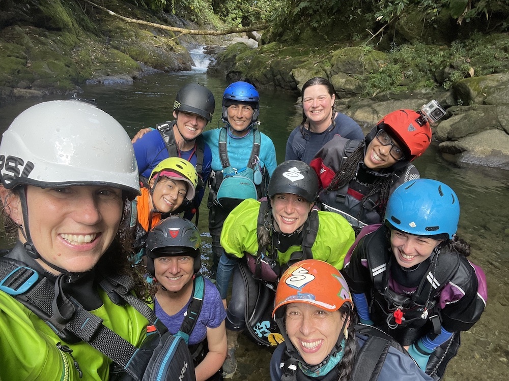 Group of women in kayaking gear posing for a group selfie in front of a creek. 