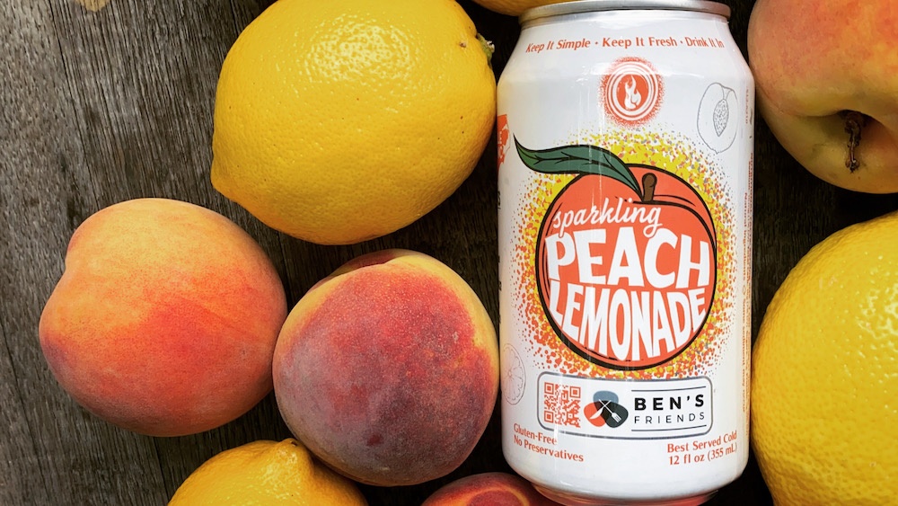 A can of Devil's Foot sparkling peach lemonade lying in the middle of lemons and peaches on a wooden table.