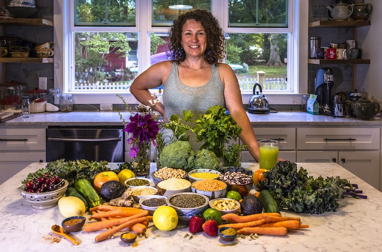 Anna Levesque, a woman, standing in a kitchen with a spread of healthy vegetable, grains, and fruits in front of her. 