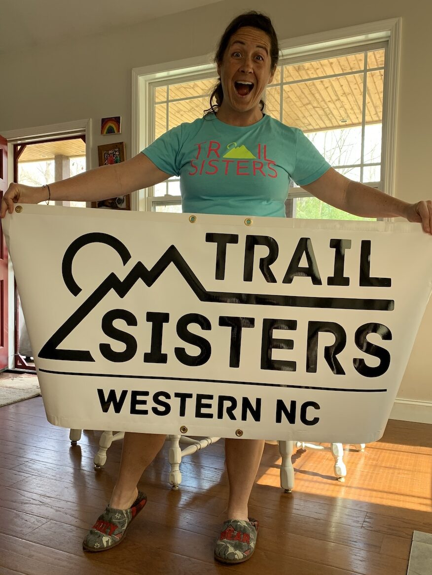 Mindy Smith holding a Trail Sisters banner that says Trail Sisters Western NC
