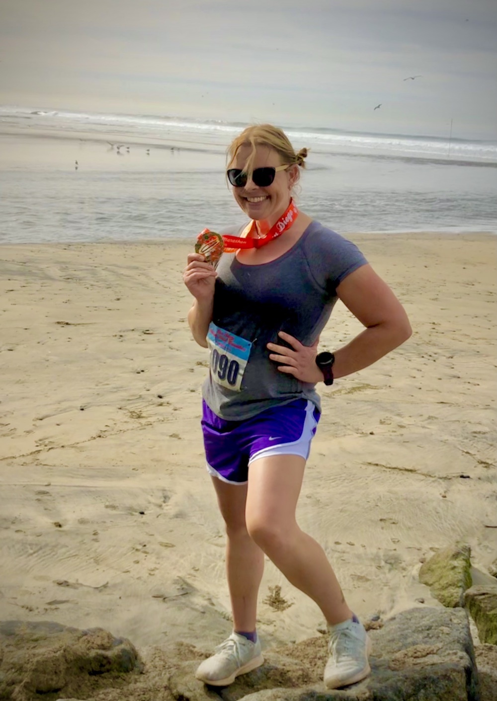 Person on beach after a running race with medal in hand and around neck. Ocean behind her.
