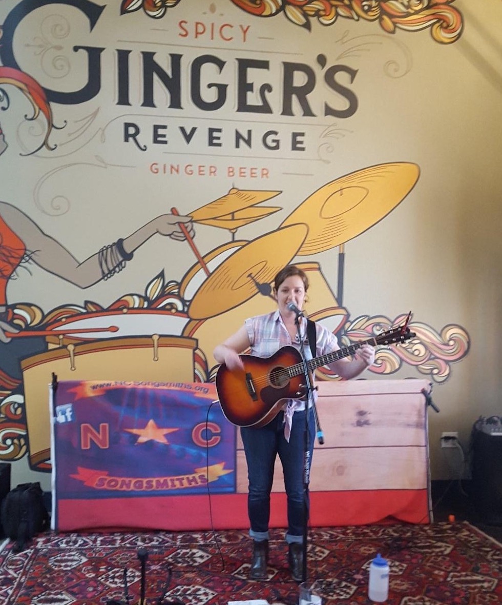 Woman singing and playing guitar in front of flag saying NC Songsmiths and Ginger's Revenge Ginger Beer Sign
