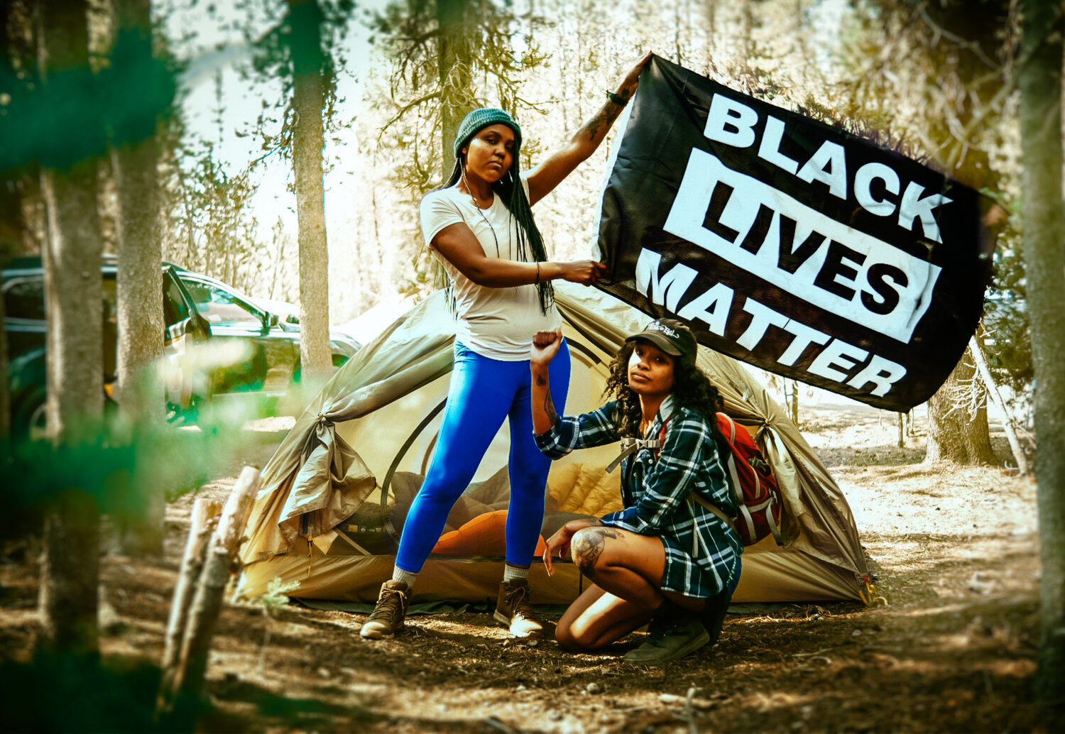 Two women standing in the woods in front of a tent holding a Black Lives Matter flag