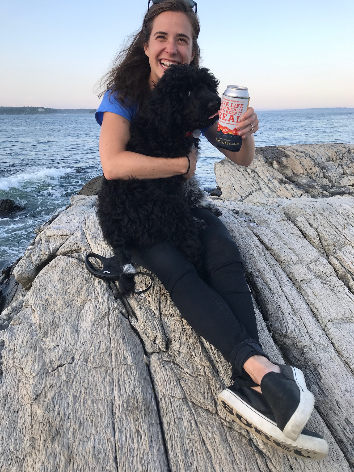 Woman sitting on the coast of Maine with a beer in hand and dog in lap