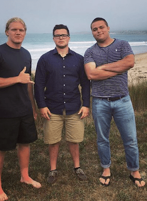 3 guys standing together on a beach with the ocean behind them. 
