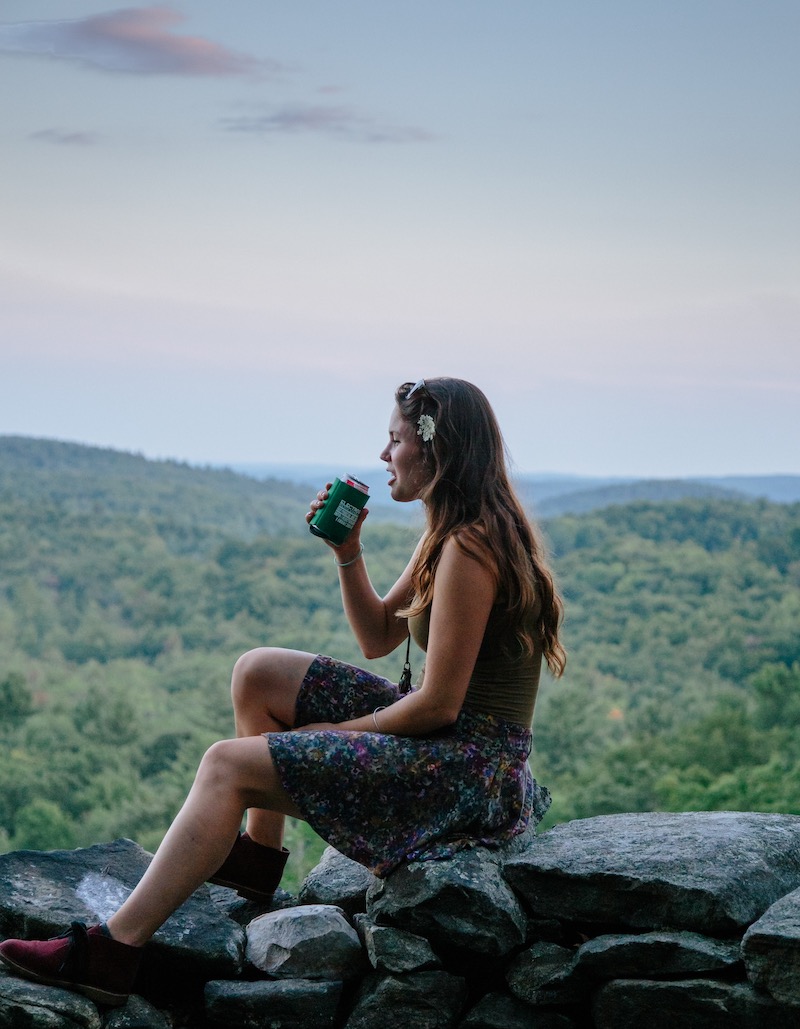 Woman with brown hair drinking beer on rock wall with mountains and green trees in background. 