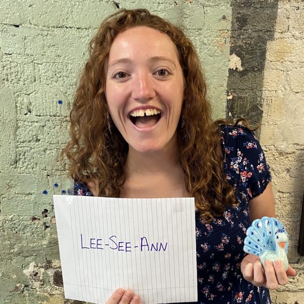 Woman against brick wall holding a ceramic peacock and a piece of paper phonetically spelling out her name "Lee-See-Ann"