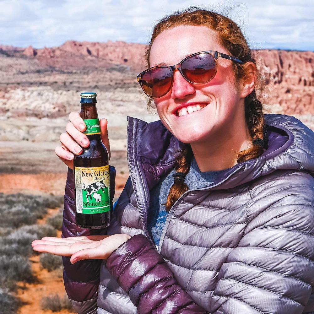 Woman standing in the desert in a puffy jacket holding a Spotted Cow beer. 
