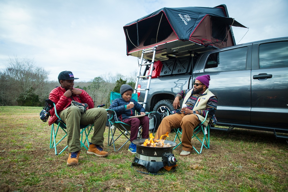 A family sitting around a fire with an overlanding rig behind them
