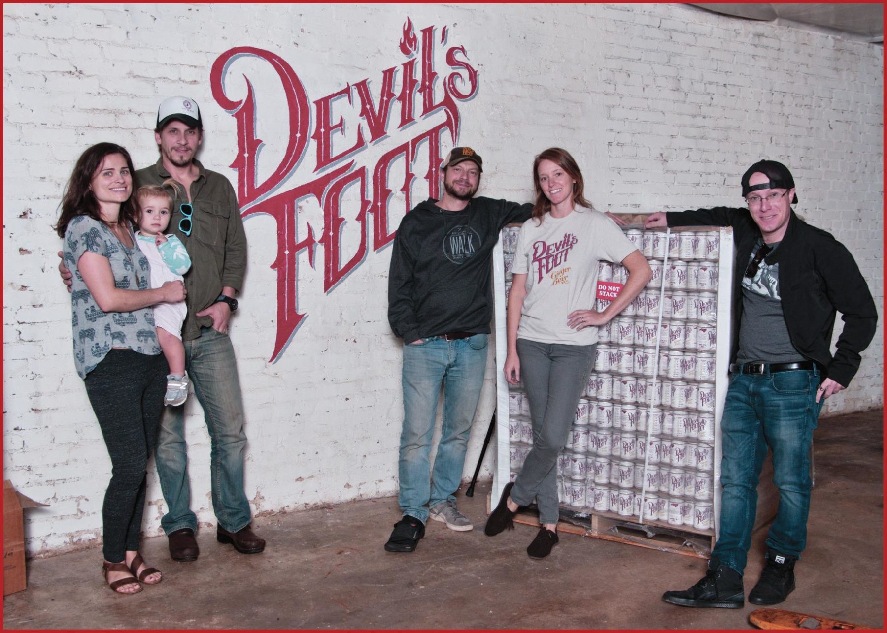 A group of people standing in a warehouse with the word's Devil's Foot spraypainted on the wall and a pallat of ginger beer cans. 