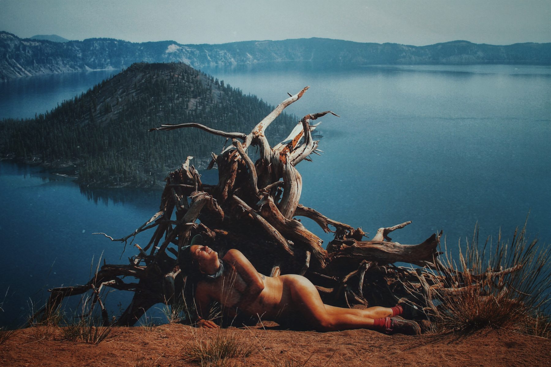 A woman lies in front of a tangled piece of wood in front of a lake surrounded by low mountains. 