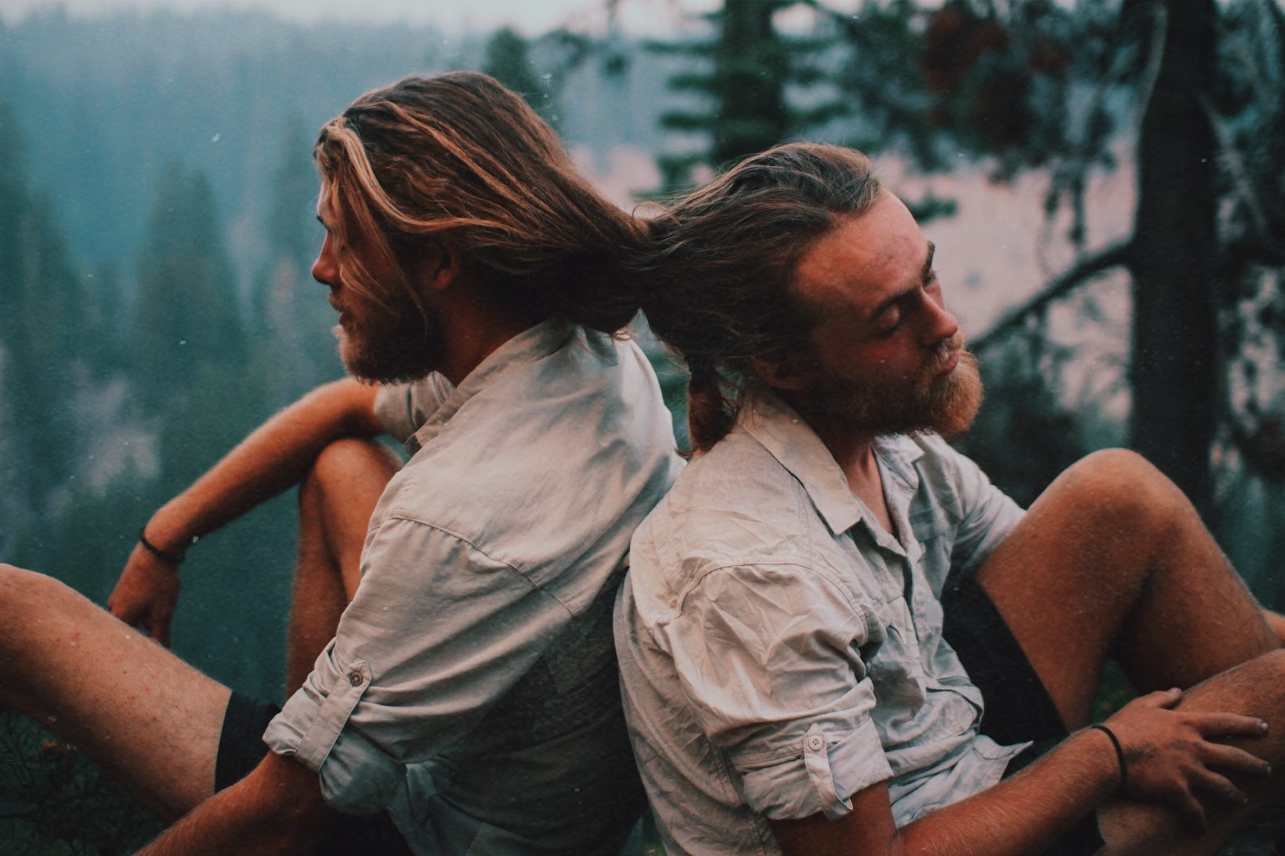 Two men in matching button-up shirts sitting in the woods with their hair braided together. 