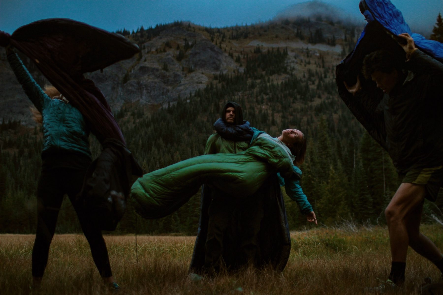 People in sleeping bags dancing and posing in a fashion shoot style but they are in a field with rocky mountains behind them. 