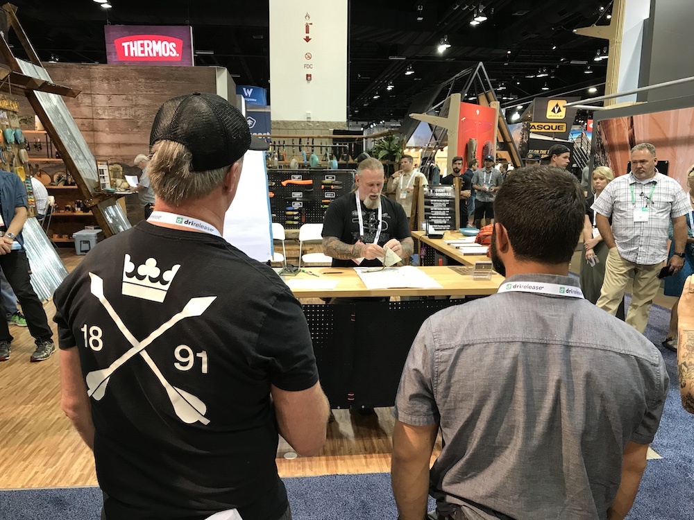 two people with backs to the camera watching another man doing a presentation at an exhibit booth.People walking by looking at man in booth. 
 