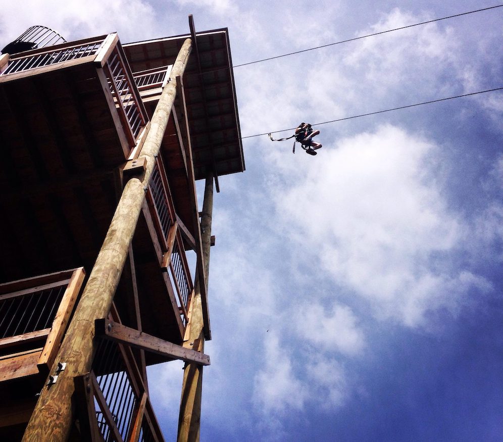 Looking up at a person ziplining with blue sky and clouds above them. 